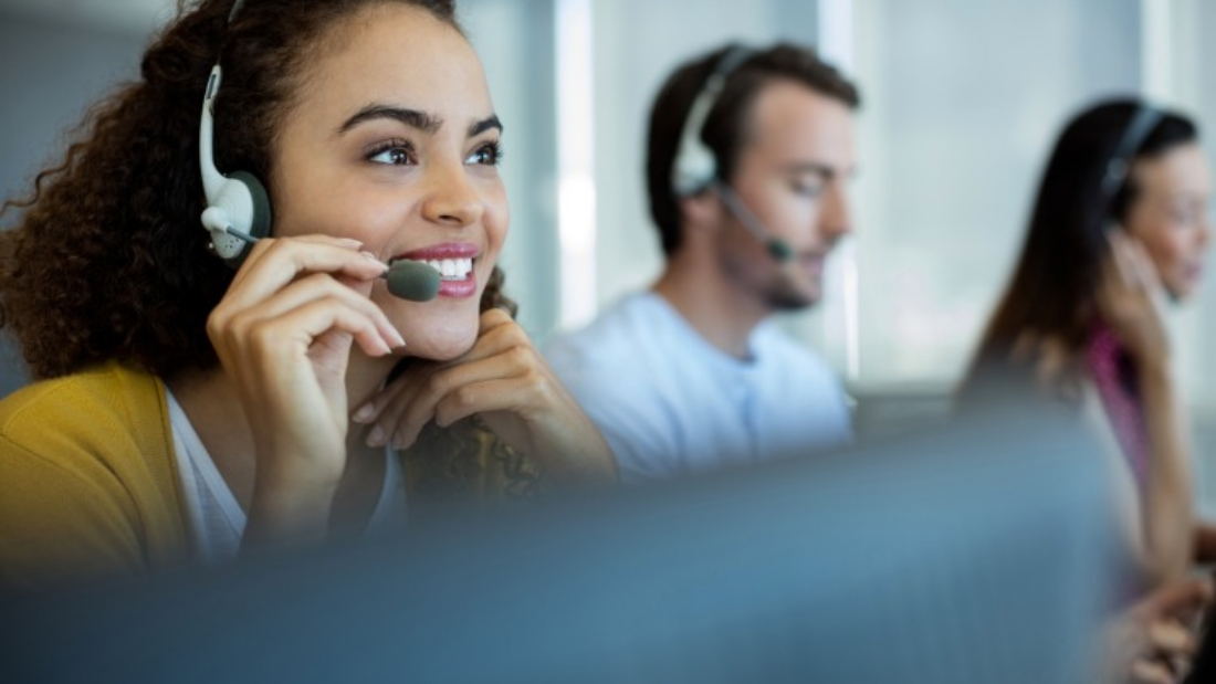 Woman sits at desk with earpiece on smiling and talking into phone