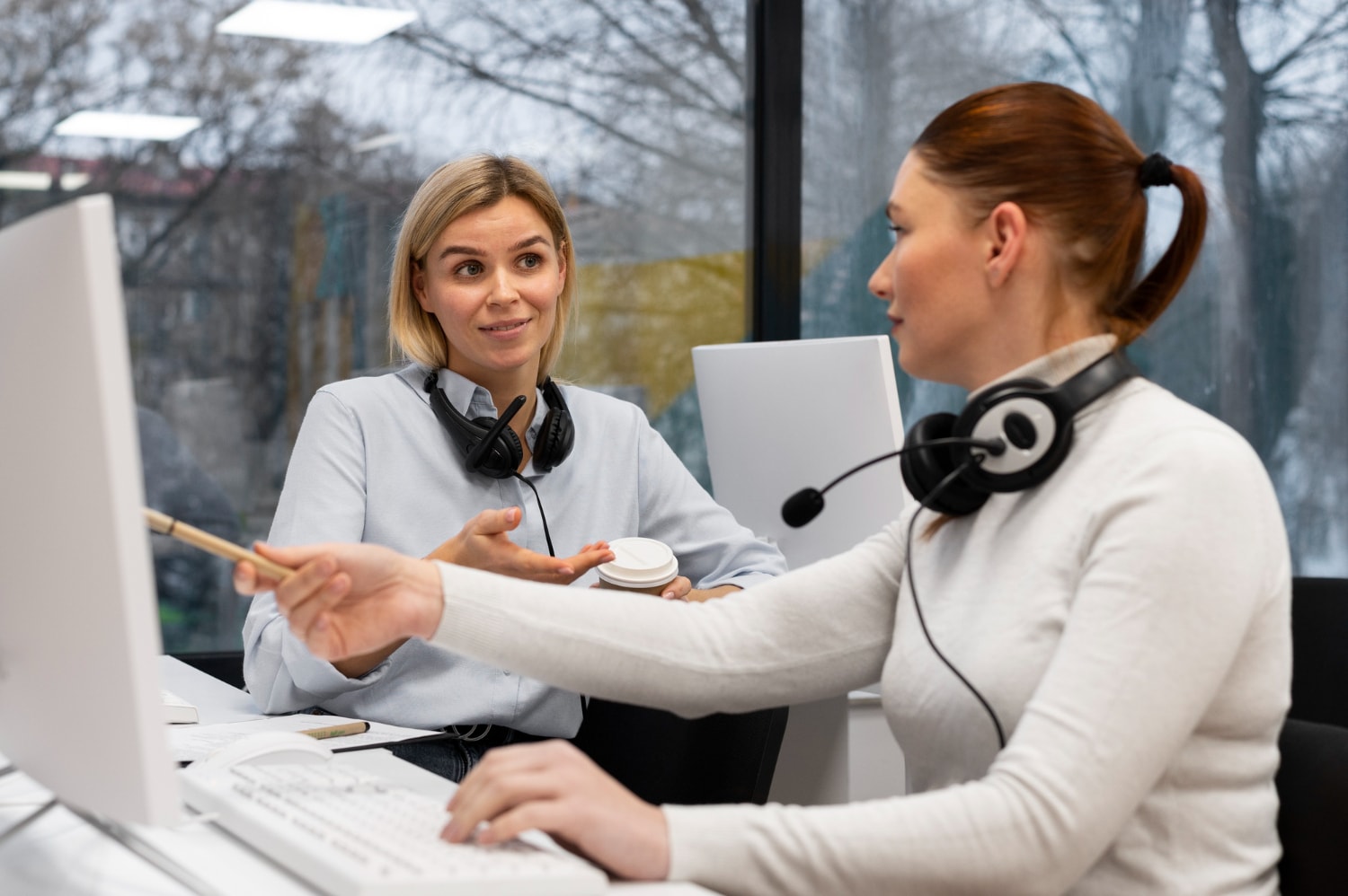 Two female customer support representatives in a modern office collaborate on a task while using a soft phone system.