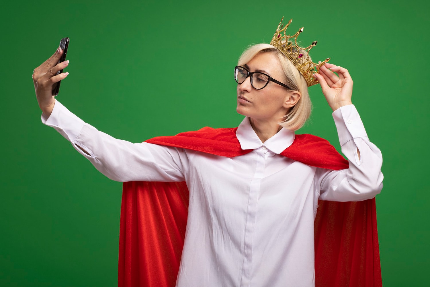 A confident woman wearing a gold crown and a red cape takes a selfie with her smartphone against a green background. She adjusts her crown with one hand while holding the phone with the other, striking a regal pose.