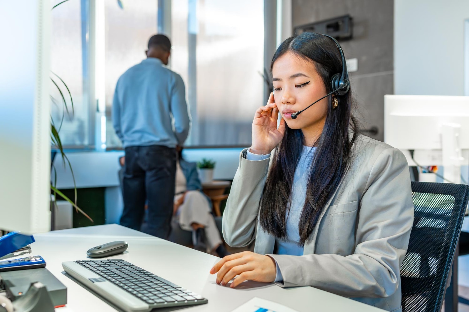 A professional woman wearing a headset sits at her desk in a modern office, engaged in a call using VoIP features.