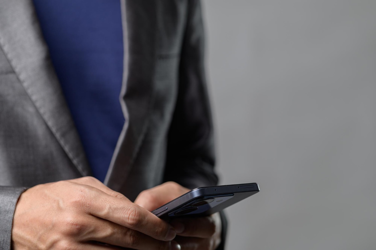 A close-up of a professional wearing a gray blazer and a navy blue shirt, holding a smartphone. The image suggests the use of VoIP features for business communication, with call routing and voicemail-to-email enabling seamless connectivity.