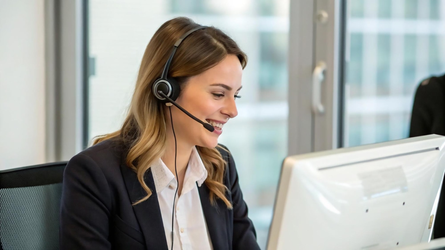 A smiling customer service representative wearing a headset is engaged in a call at her workstation in a modern office. The presence of VoIP features enables efficient communication, with call routing ensuring seamless connections and voicemail-to-email allowing for easy message retrieval. 