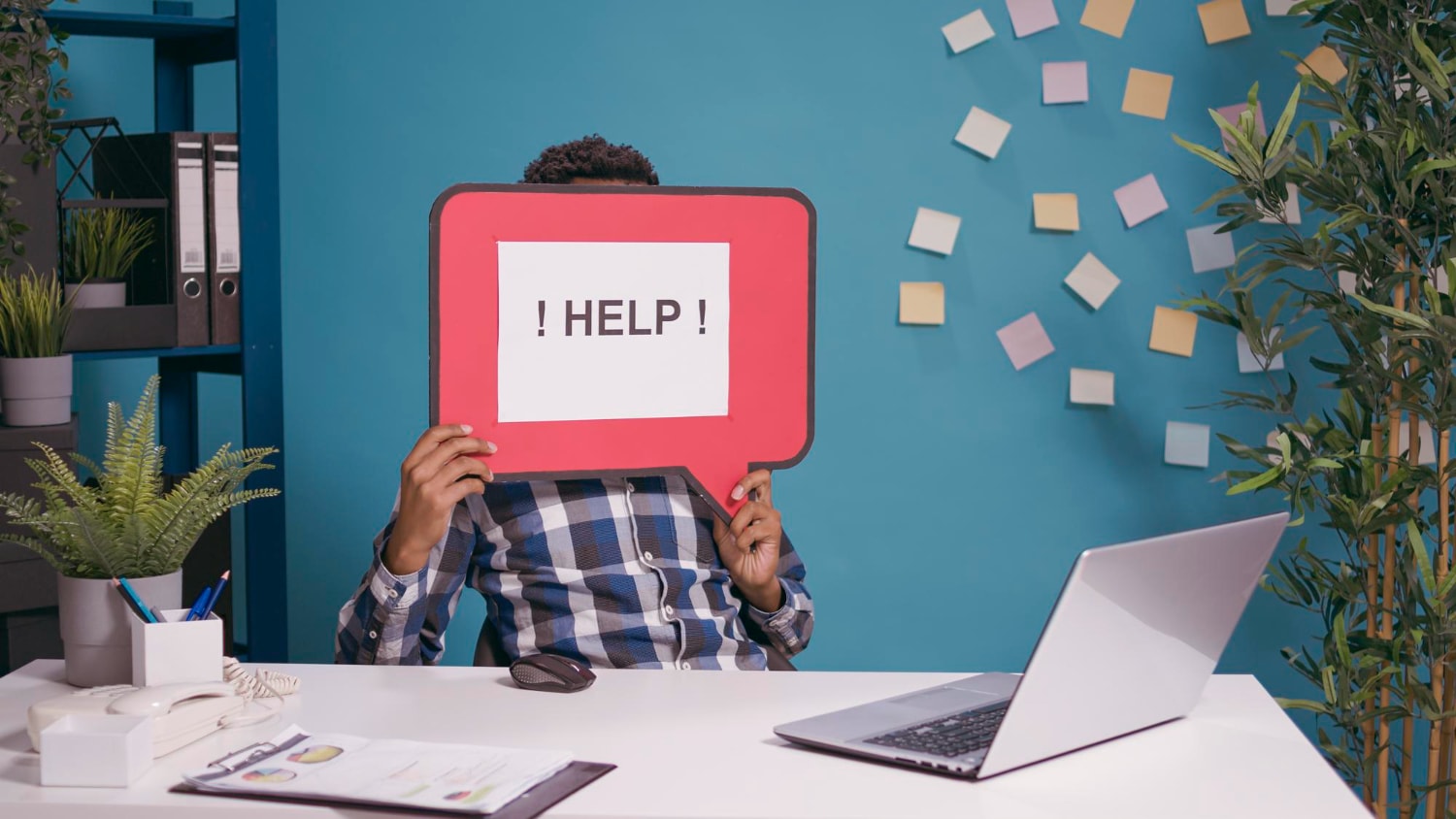 A frustrated office worker sits at a desk holding up a large red speech bubble sign with the words "! HELP !" written on it, covering their face. The modern workspace features a laptop, documents, a landline phone, and a neatly organized background with sticky notes on a blue wall illustrating workplace communication struggles and the need for scalable communication solutions.