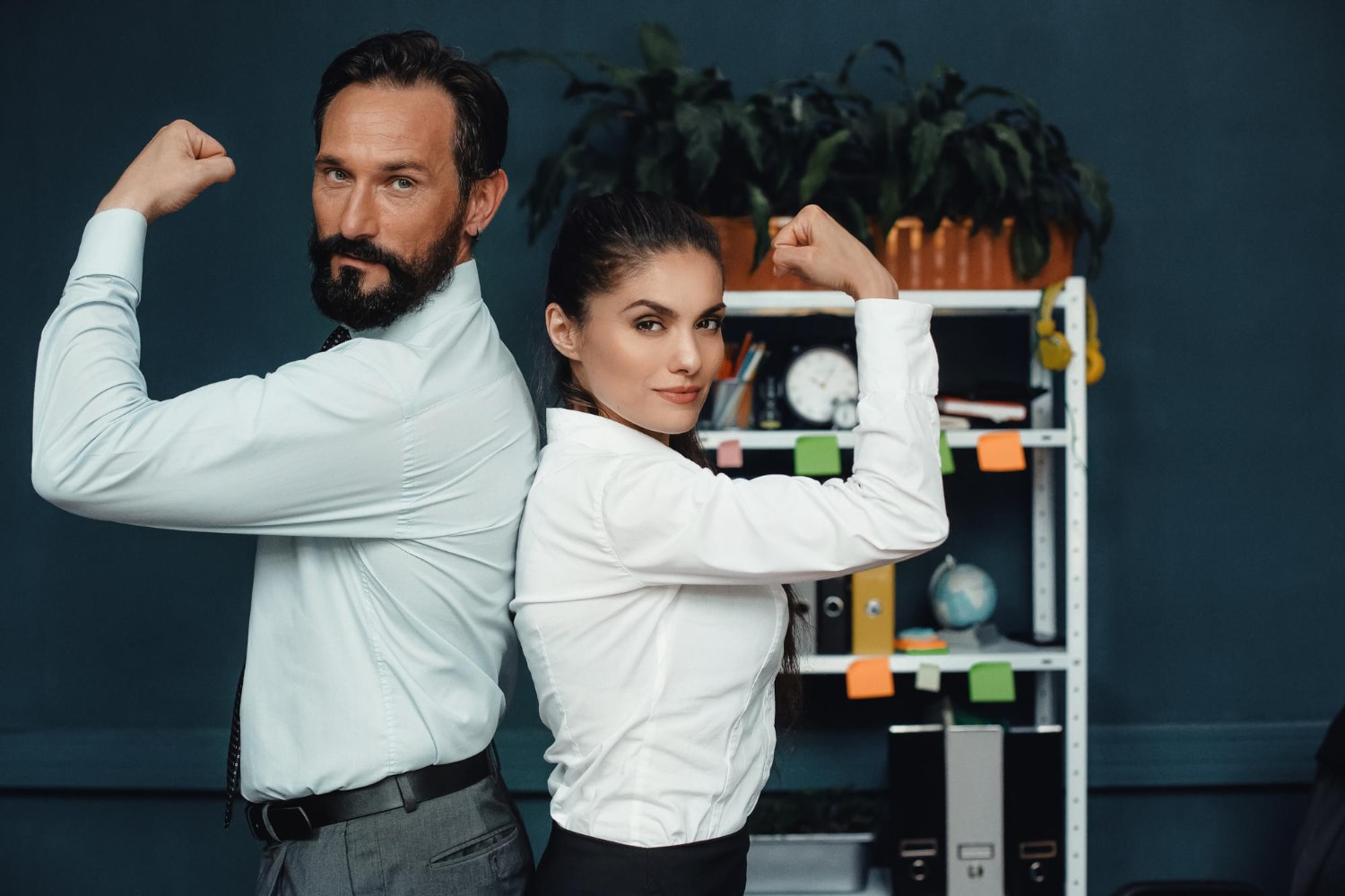 A confident male and female business team stand back-to-back in an office, flexing their arms in a show of strength and teamwork. Both are dressed in professional attire, exuding determination and collaboration. Behind them, a well-organized office space with shelves, documents, and sticky notes represents productivity and efficiency symbolizing business connectivity.