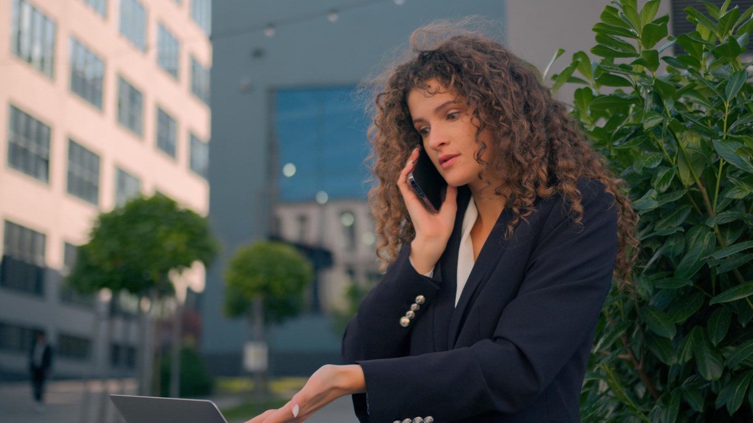 A professional woman with curly hair, dressed in a dark blazer, is standing outdoors in a business district while multitasking. She is engaged in a phone call using VoIP features like call routing, while also working on her laptop, possibly accessing voicemail-to-email services.