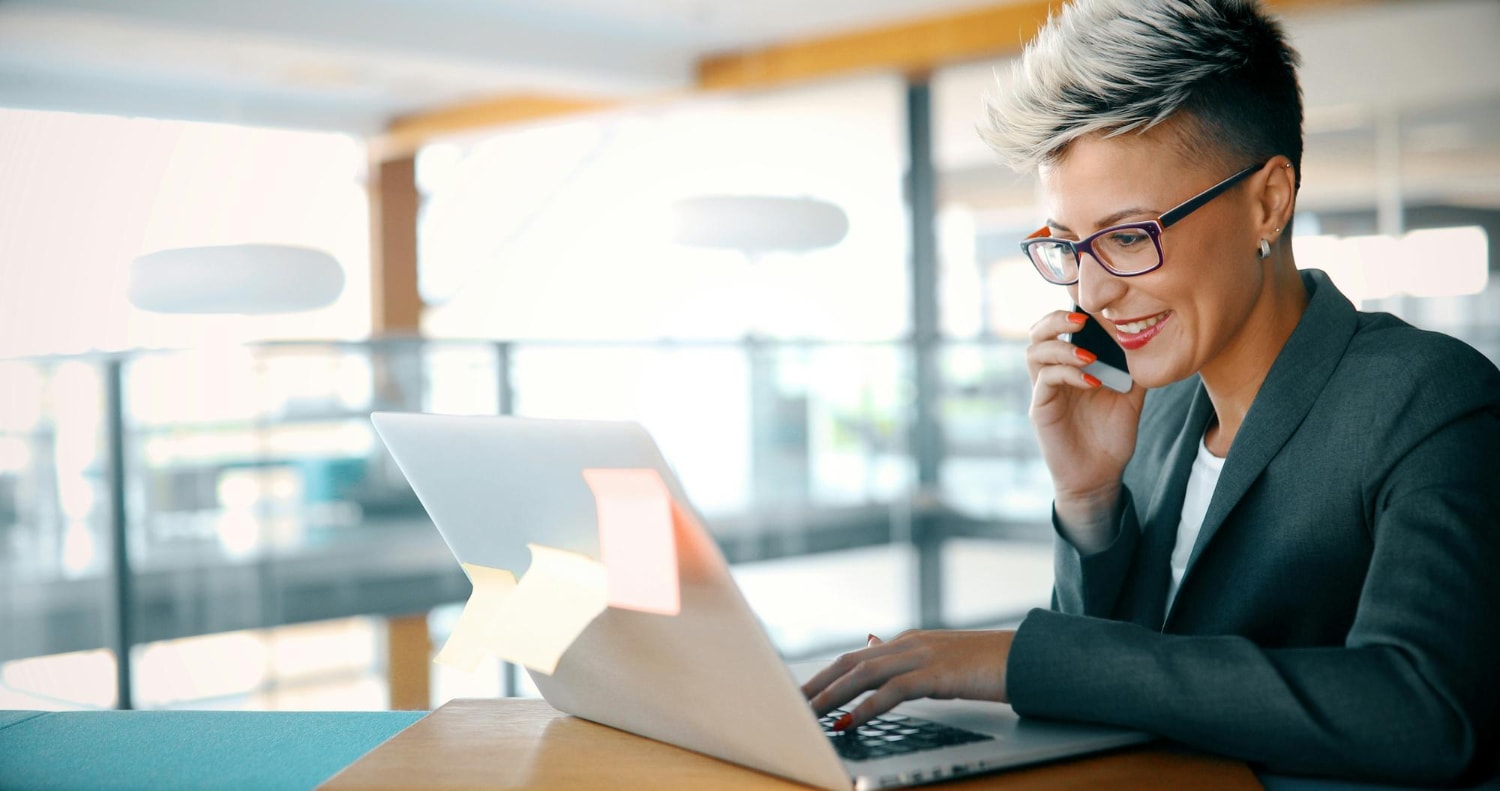 "A stylish professional with short, edgy hair and glasses is seated at a modern workspace, smiling while engaged in a phone call.
