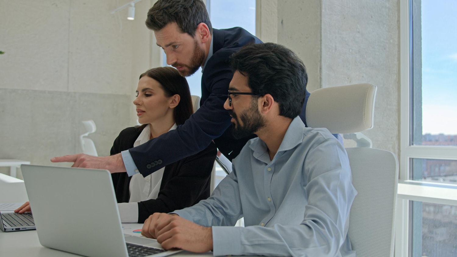 "A team of three professionals collaborates in a modern office with large windows. A man in a dark suit leans over, pointing at a laptop screen, while his colleagues, a woman in a blazer and a man in a light blue shirt, engage in the discussion. Their teamwork suggests the use of collaboration tools.