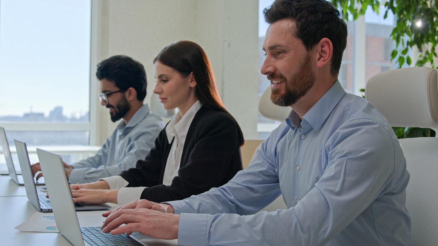 A group of three professionals sits in a modern office space, working on laptops with a focus on collaboration.