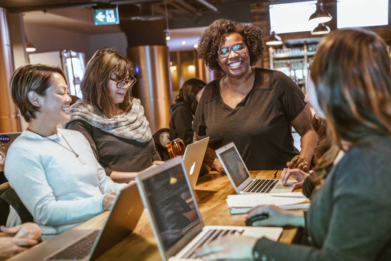 A diverse group of professionals gathers around a wooden table in a cozy, modern workspace, engaged in a collaborative discussion. Laptops are open, and notebooks are being used, suggesting the integration of collaboration tools for efficient teamwork.