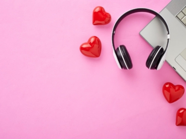 A modern workspace setup featuring a laptop, a pair of black and white headphones, and several glossy red heart-shaped decorations on a pastel pink background. Representing Soft phones