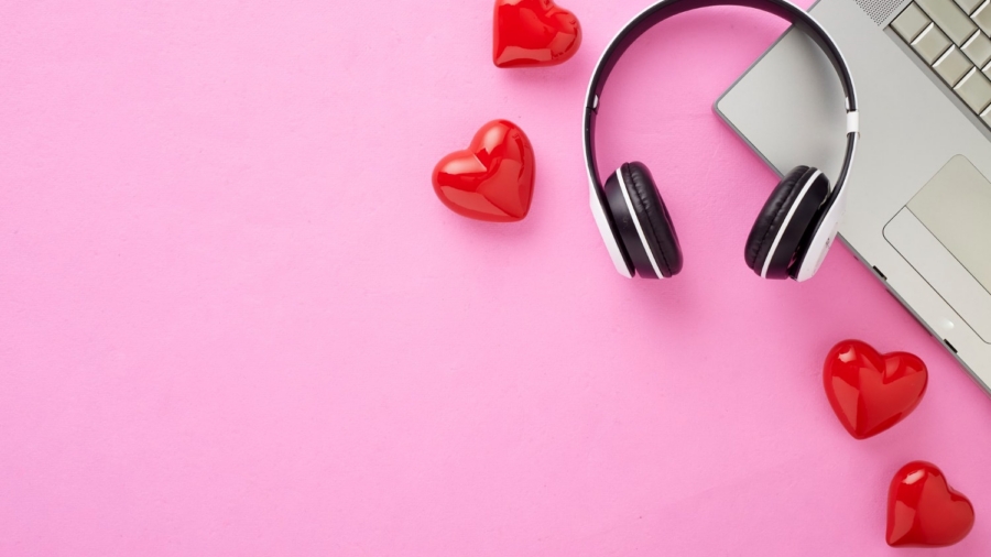 A modern workspace setup featuring a laptop, a pair of black and white headphones, and several glossy red heart-shaped decorations on a pastel pink background. Representing Soft phones