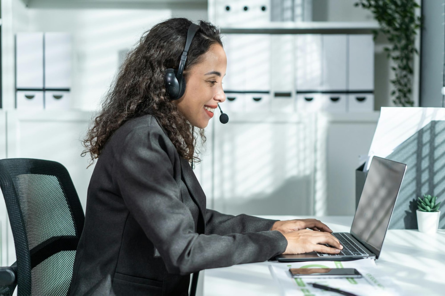 A customer service representative wearing a headset sits at a desk in a modern office, smiling while working on a laptop suggesting the use of VoIP enhances seamless communication.
