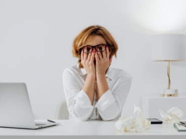 Woman with hands on face, sitting at desk with laptop and tissues, feeling stressed.